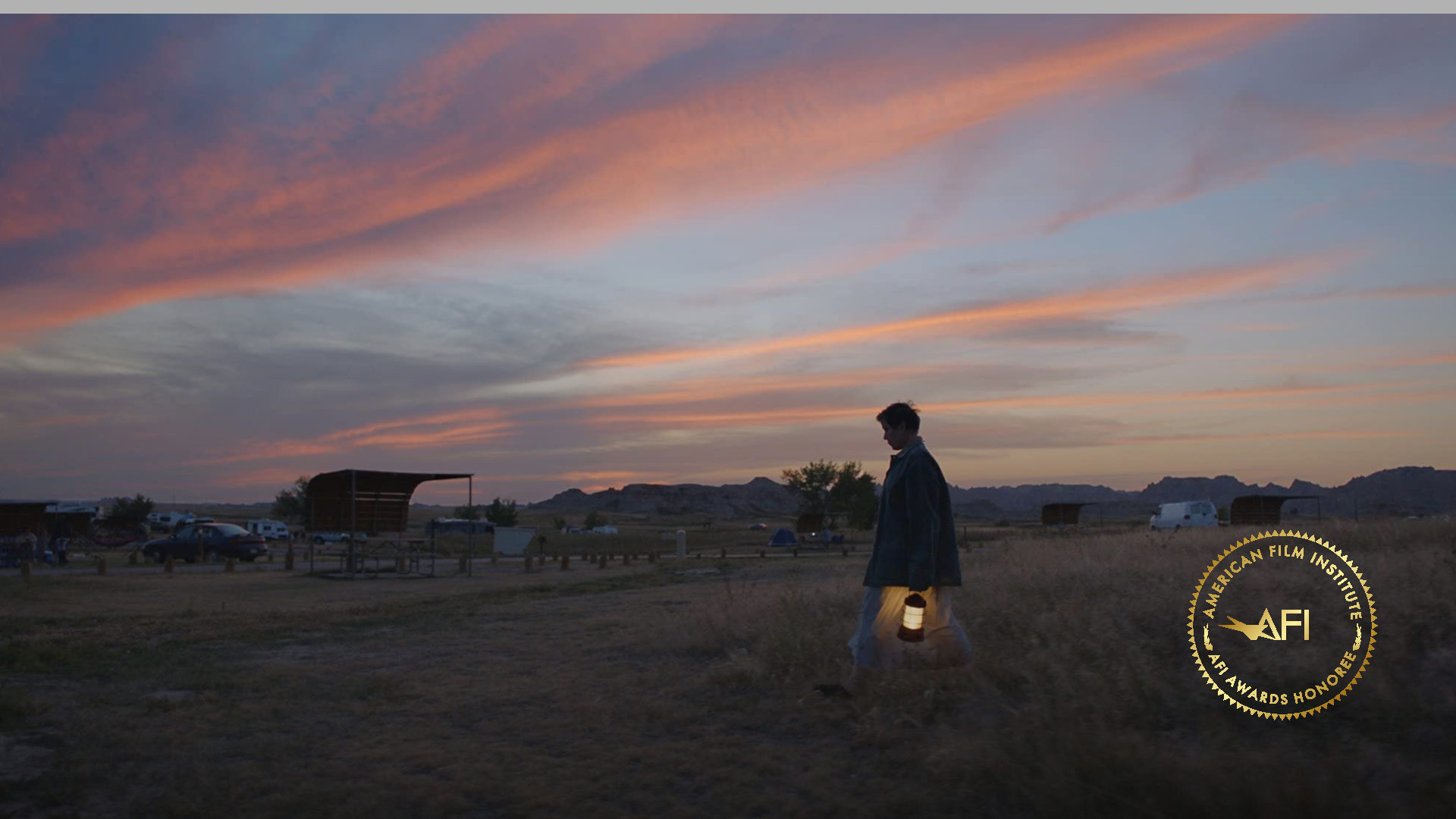 In the righthand lower corner is the AFI AWARDS logo. The image is a film still from NOMADLAND: Fern (Frances McDormand) is walking across an open field holding a lantern. The clouds, set against a blue-ish gray sky, are a tinged dark orange from the setting sun.