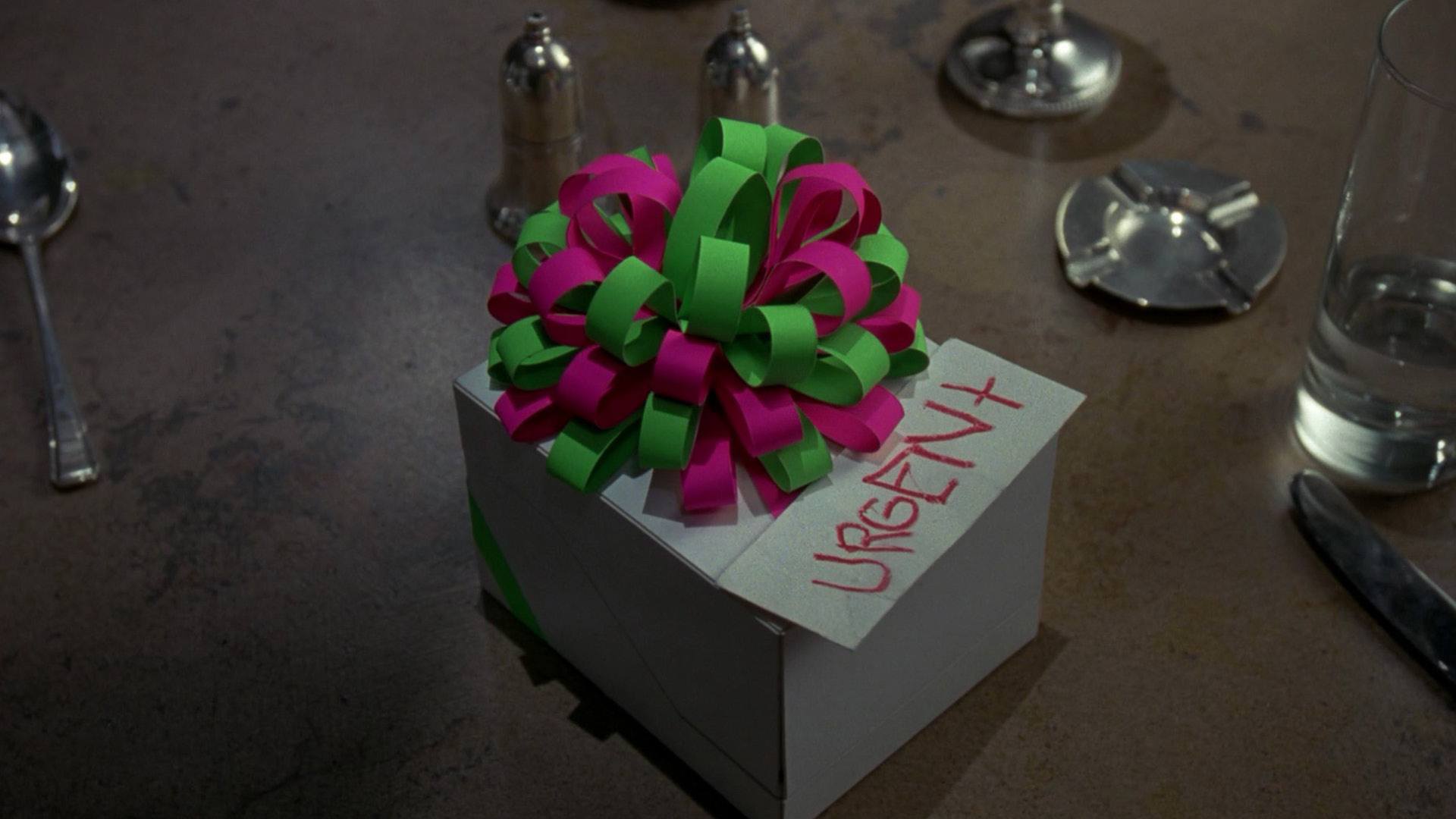 BATMAN film still of a white package with a pink and green bow and a note with the word "URGENT" written on it.