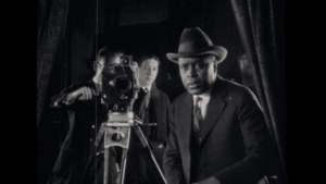 Black and white image of American film director Oscar Micheaux (1884-1951) in a suit and hat standing next to a film camera -Photo Credit - Kino Lorber